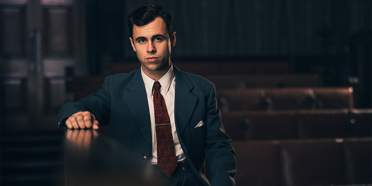 A young man in a blue suit sits confidently, resting his hand on a wooden railing in a courtroom.