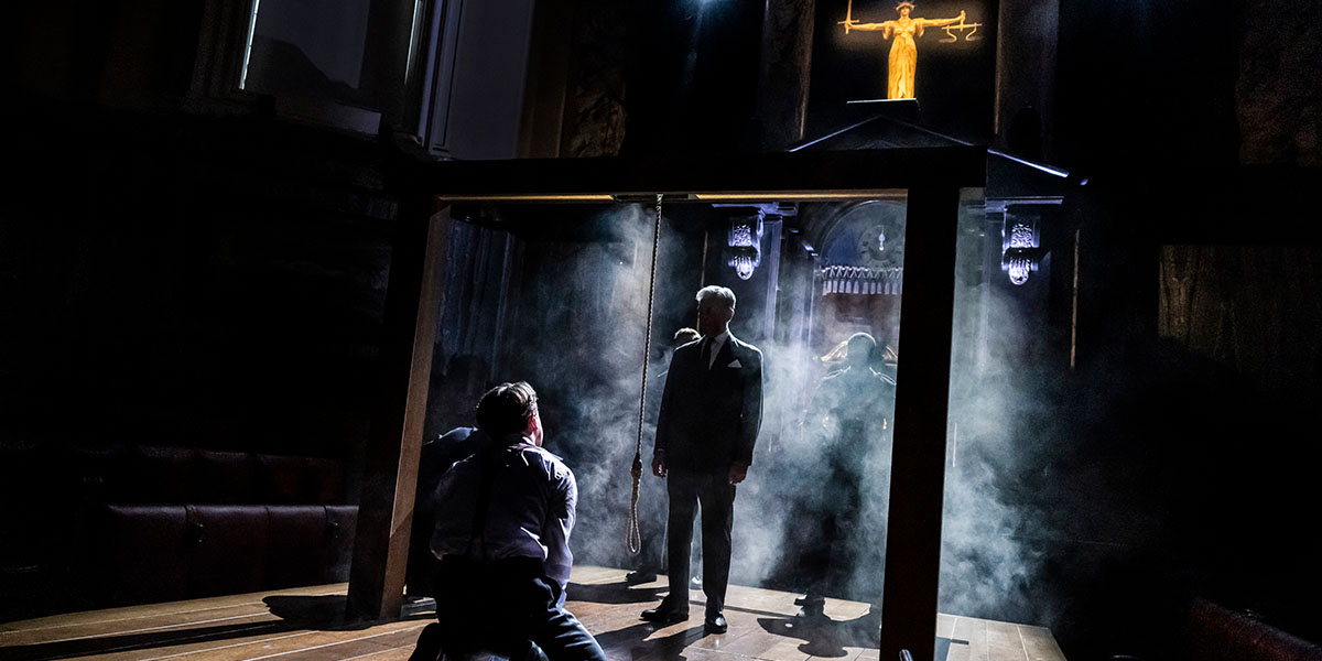 A man on his knees in front of a gallows, inside a courtroom. The lighting is dark and smoky
