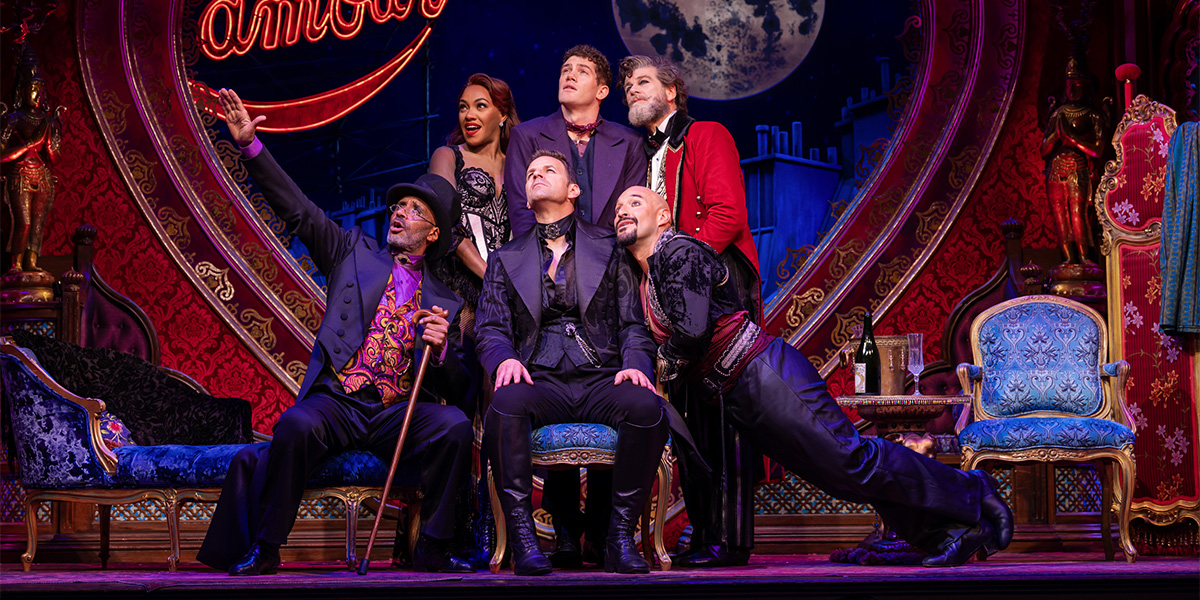 The cast of Moulin Rouge! The Musical performs energetically on stage, wearing vibrant costumes against a dramatic backdrop.