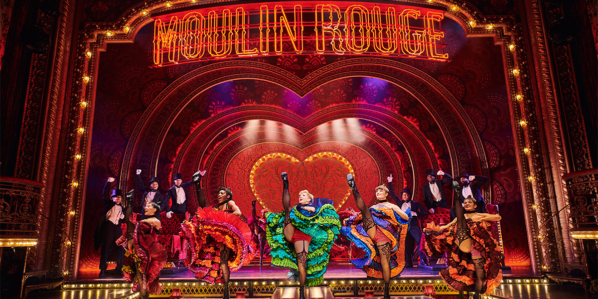 The cast of Moulin Rouge! The Musical performs energetically on stage, dressed in vibrant costumes with dramatic lighting.