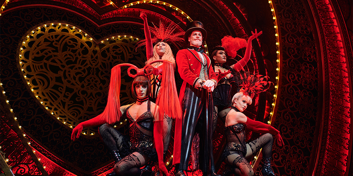 A person dressed in a red jacket and a black top hat stands surrounded by people wearing black and red corseted outfits stand on stage.