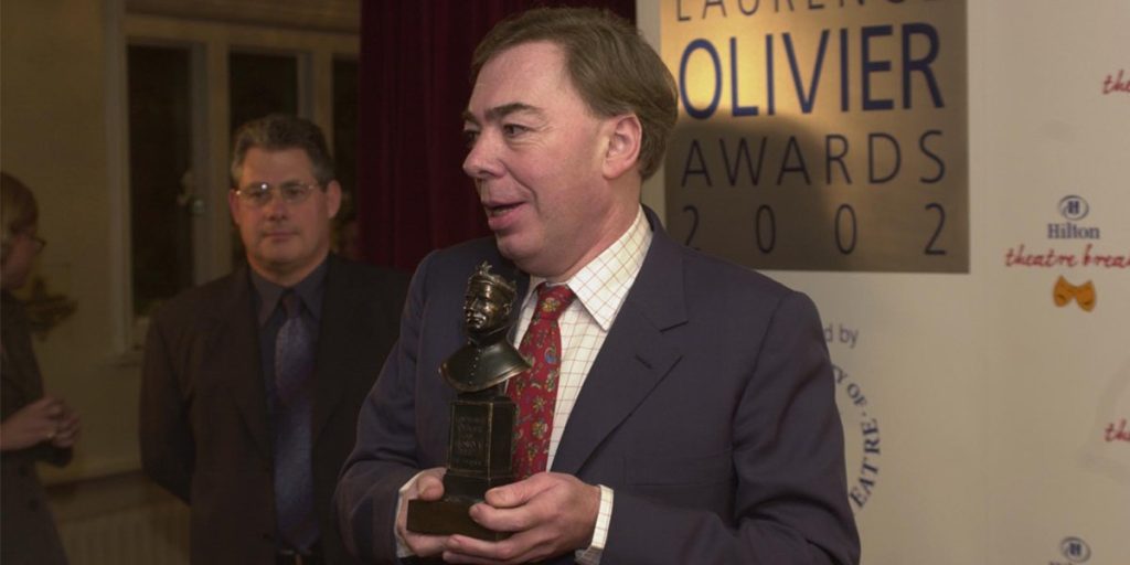 A man in a suit holds an Olivier Award trophy, smiling, with a banner in the background.