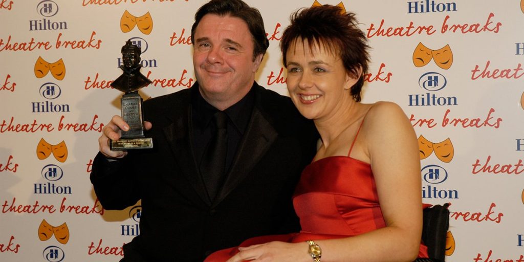 A group of elegantly dressed individuals stands together, smiling, with a red carpet and awards in the background.
