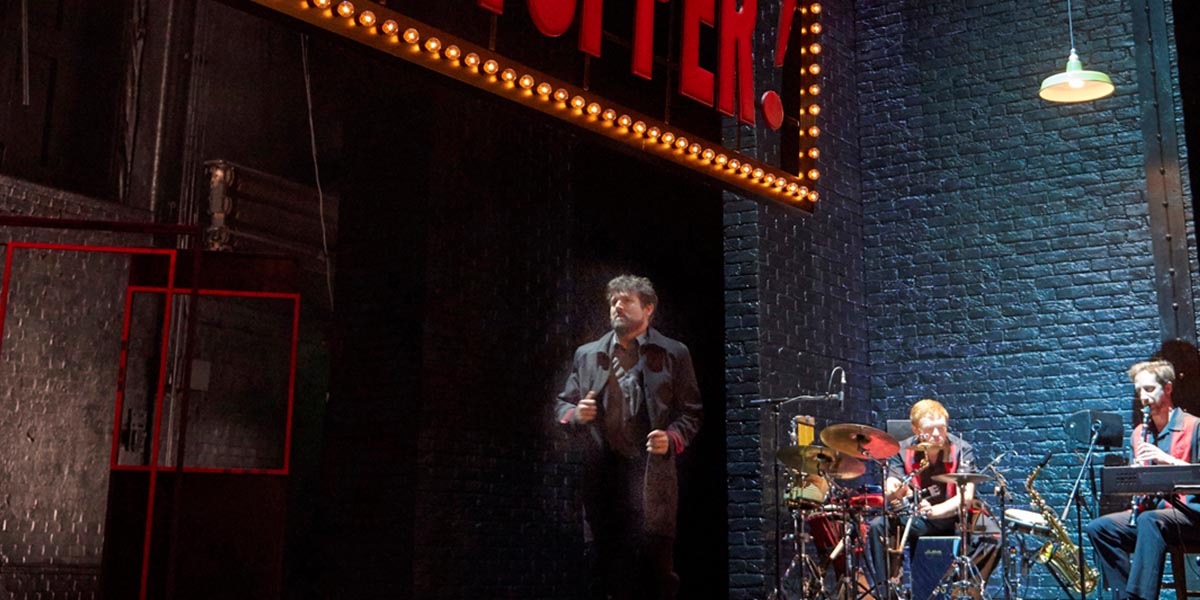 A performer in a vibrant costume gestures expressively on stage, with bright lights illuminating the backdrop.