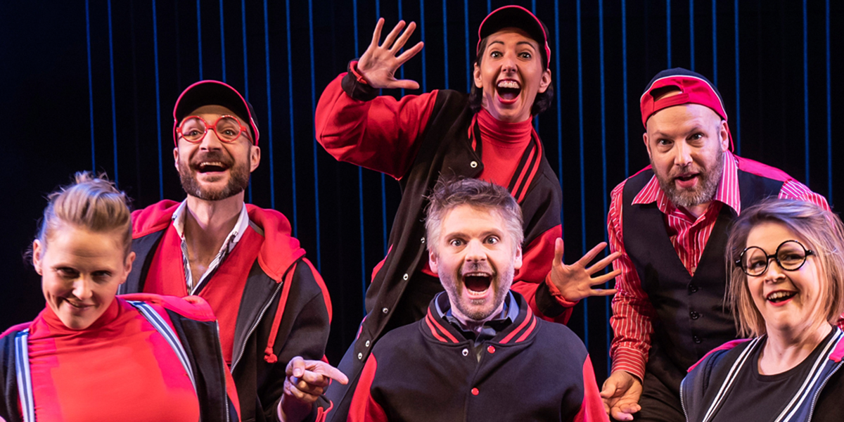 A group of performers in colourful costumes energetically act on stage, with bright lights illuminating the scene.