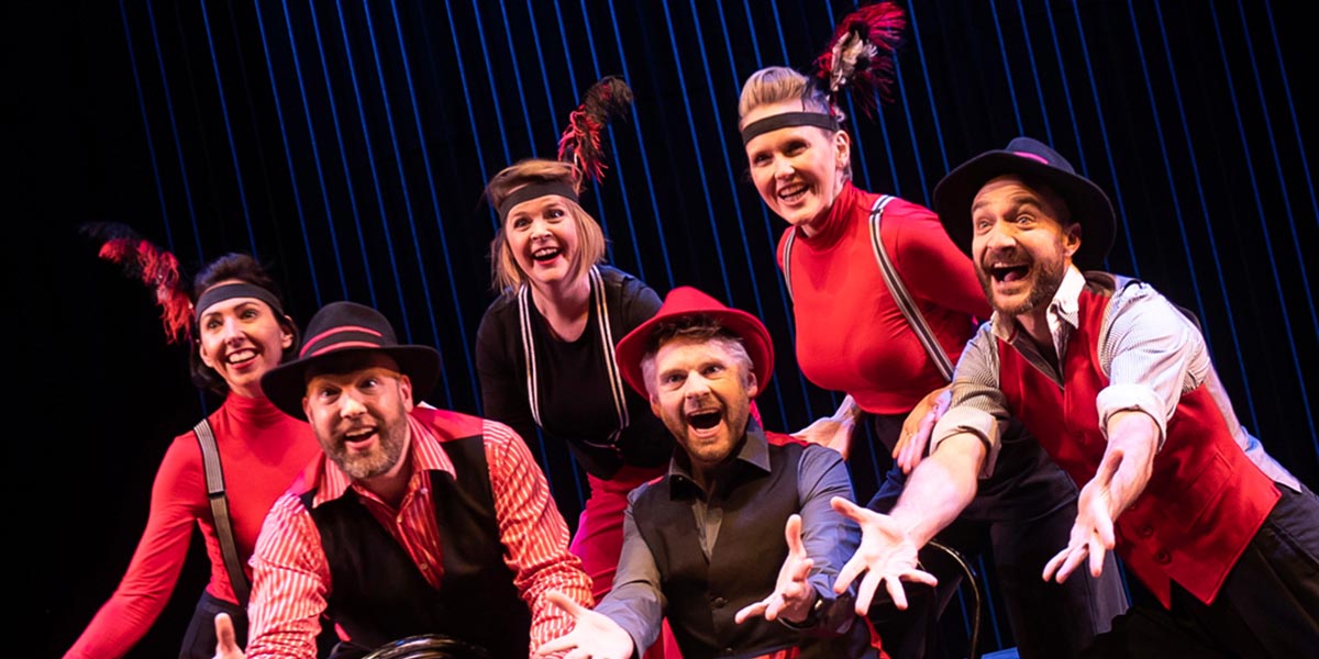 A performer in a bright costume gestures expressively on stage, surrounded by colourful lighting and audience silhouettes.