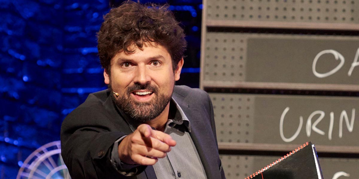 A performer in a bright costume gestures expressively on stage, with vibrant lighting and audience silhouettes in the background.