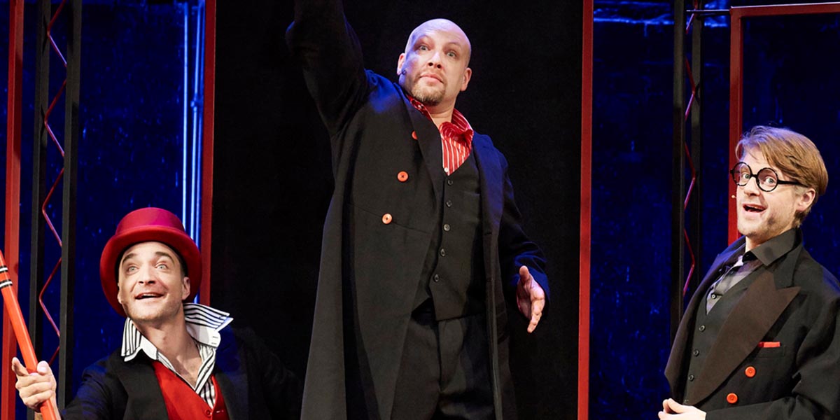 A performer in a bright costume gestures expressively on stage, surrounded by vibrant stage lights and props.