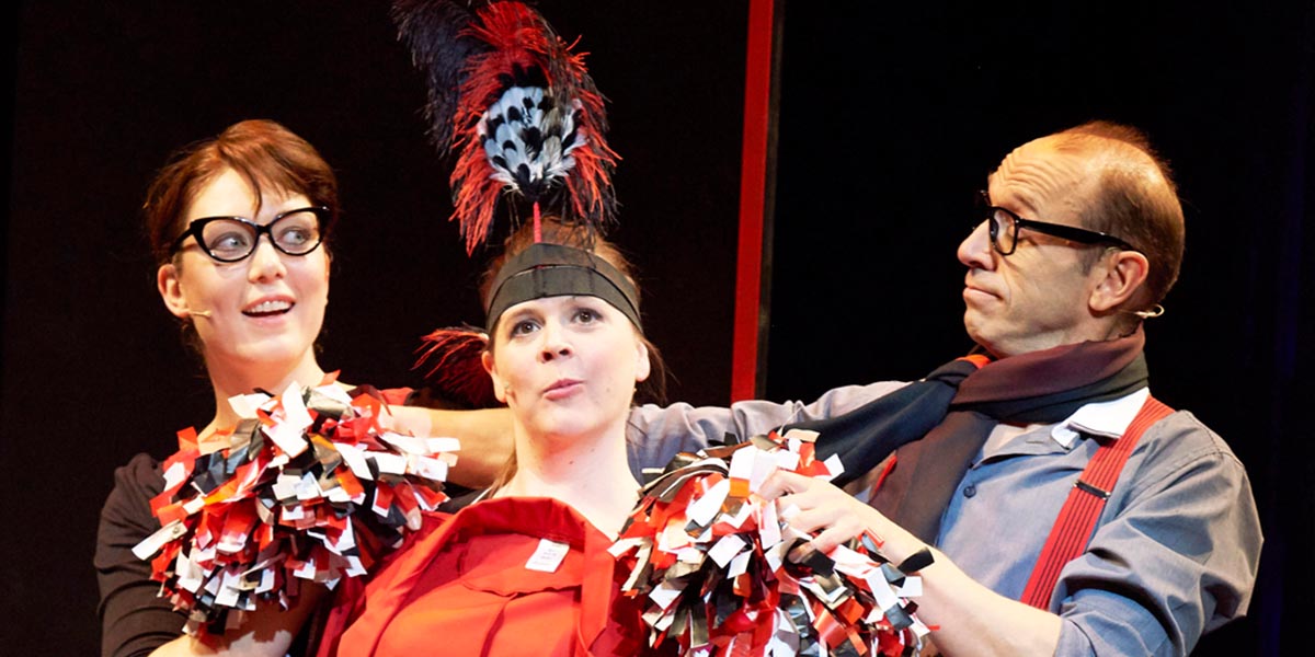 A group of performers in colourful costumes energetically act on stage, with bright lights illuminating the scene.