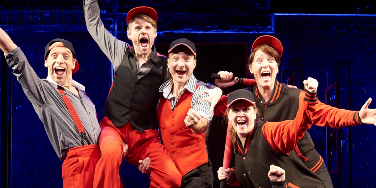 A performer in a vibrant costume gestures expressively on stage, with dramatic lighting and a cheering audience behind.