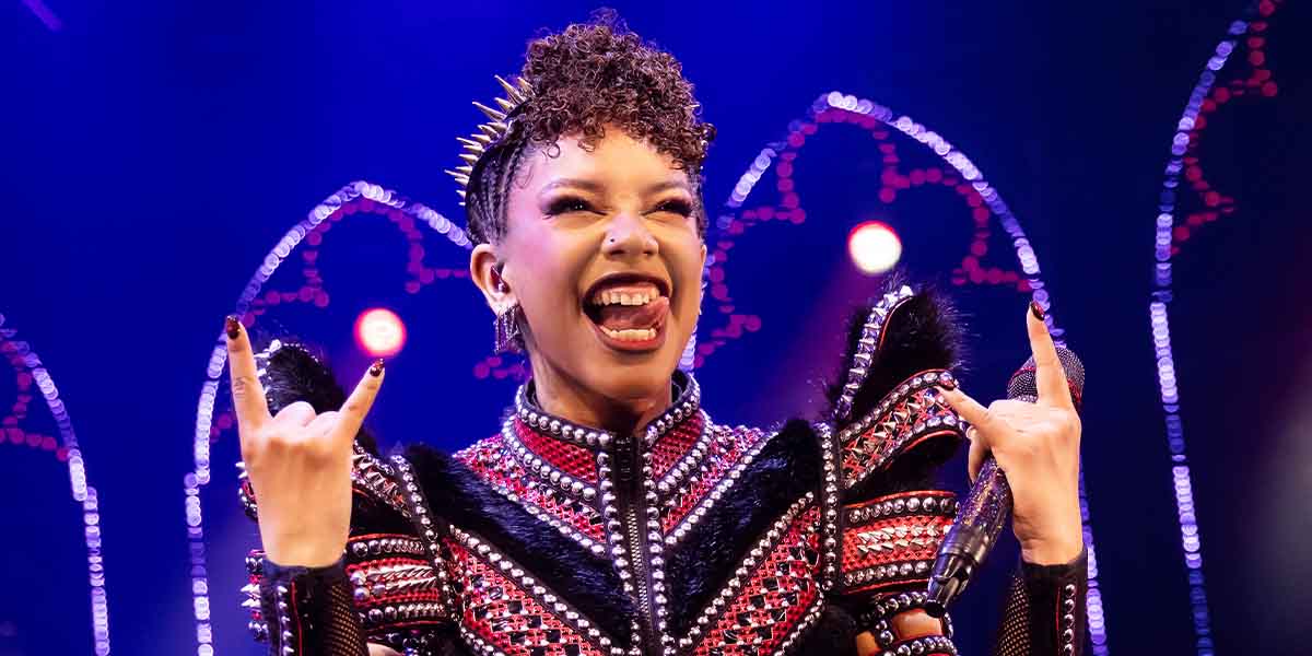 Freya Karlettis as Anna of Cleves, performing energetically with a microphone, wearing a striking embellished costume.