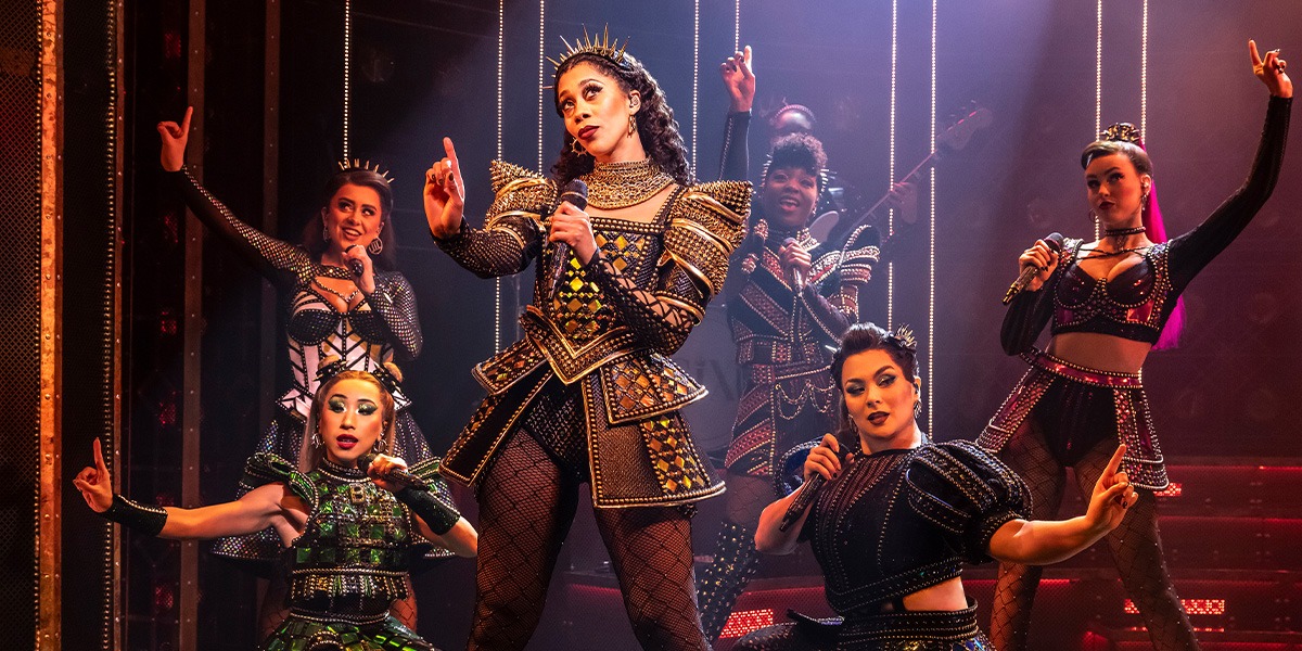Jaz Robinson as Catherine of Aragon wears a red gown, striking a confident pose against a dark background.