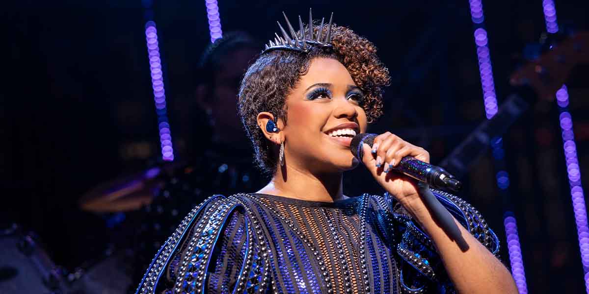 Nia Stephen as Catherine Parr, smiling and singing into a microphone, wearing a sparkling black outfit.
