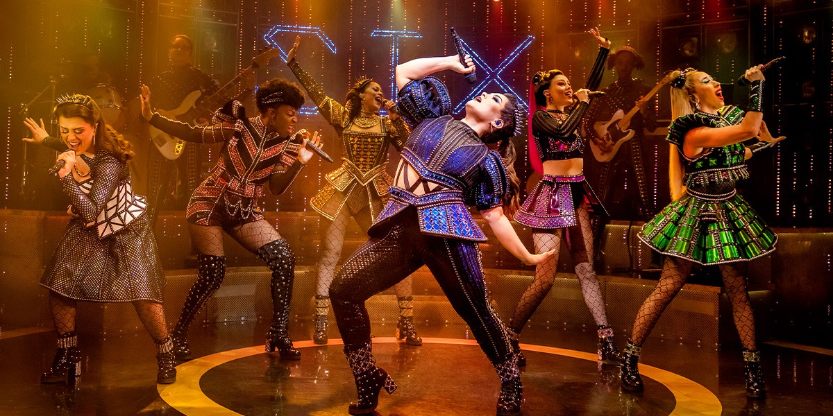 The six performers in vibrant costumes pose dynamically, showcasing diverse expressions against a dark stage backdrop.