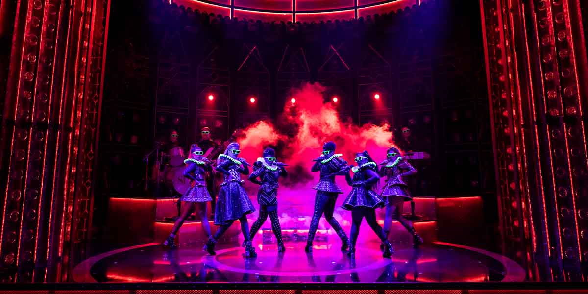 Six performers in vibrant costumes dance on stage, surrounded by dramatic lighting and smoke effects.