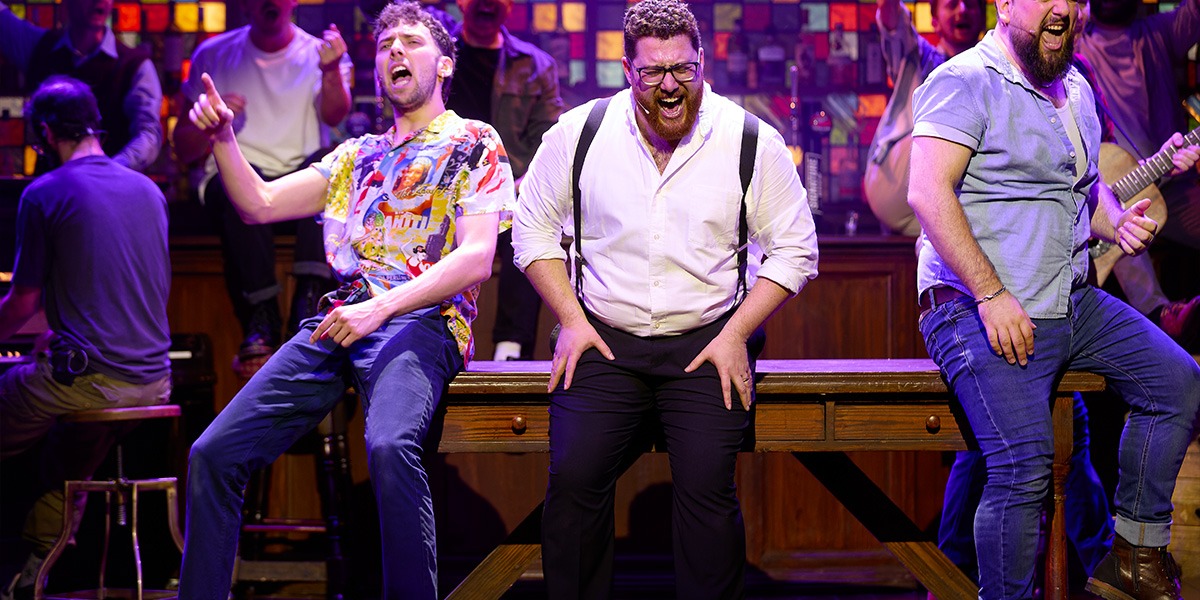 A diverse group of male singers in formal attire perform energetically on stage, with vibrant lighting behind them.