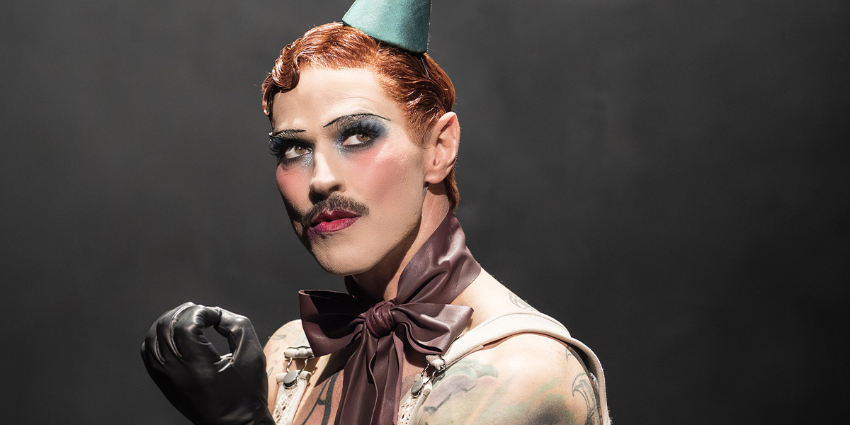 Matt Willis, as the Emcee, poses with a dramatic expression, wearing makeup, a bow tie, and a green hat.