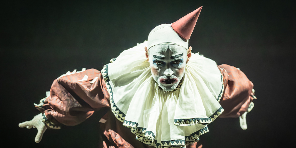 Matt Willis, dressed in a clown costume, stands on stage as the emcee, engaging the audience.
