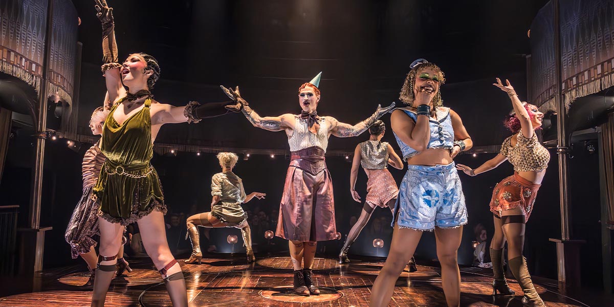 Matt Willis, as Emcee, stands centre stage in a black suit, surrounded by performers in vibrant costumes.