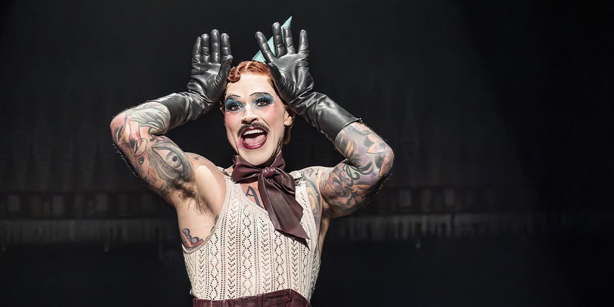Matt Willis, as Emcee, performs on stage in a tuxedo, with dramatic lighting and a vibrant backdrop.