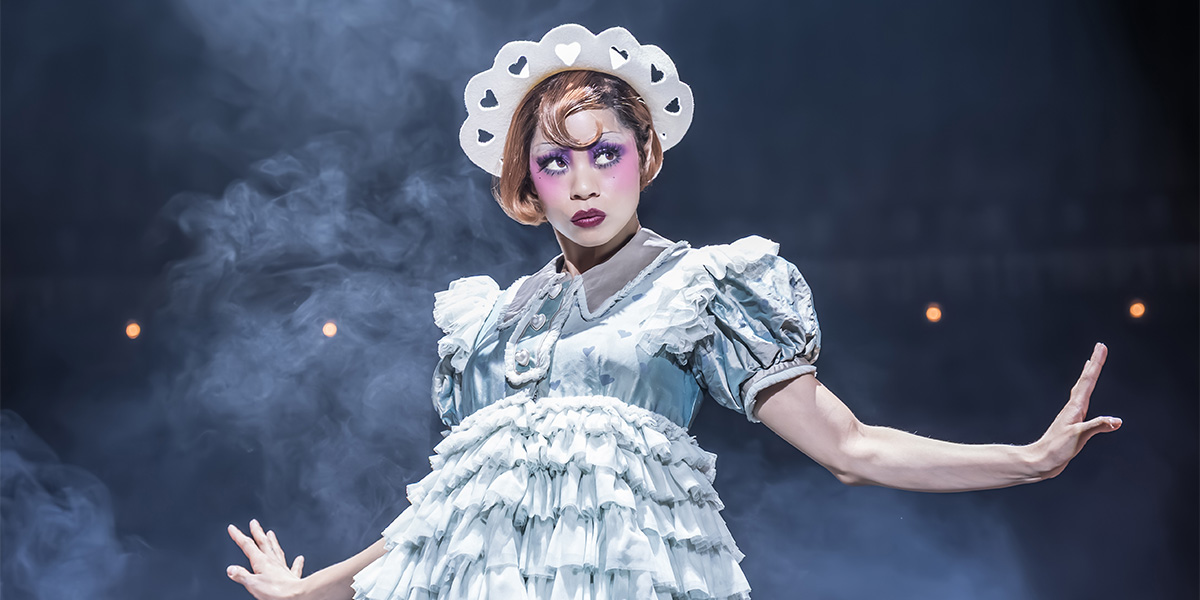 A performer in a sequined costume poses dramatically on stage, with vibrant stage lights illuminating the scene.