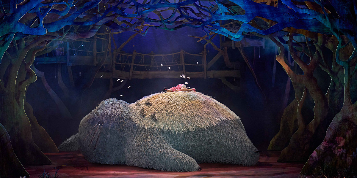 A large, grey Totoro character stands in a lush green forest, surrounded by vibrant foliage and sunlight.