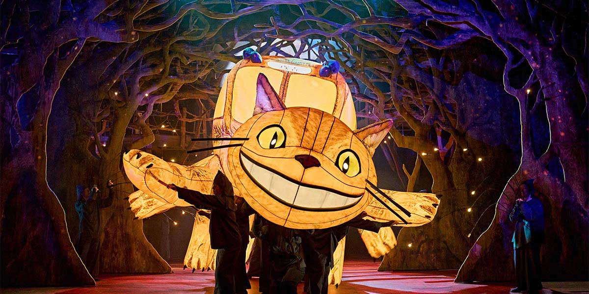 A large, fluffy grey creature with big eyes sits on a grassy hill, surrounded by trees and flowers.