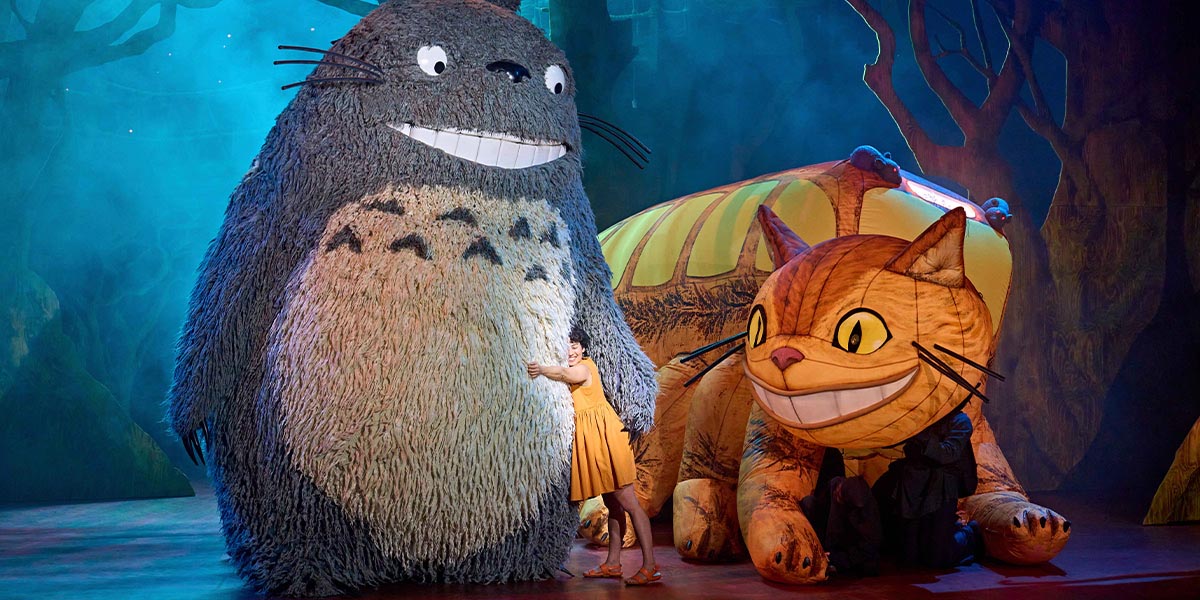 A large, fluffy grey creature with big eyes sits among lush green foliage, surrounded by vibrant flowers.