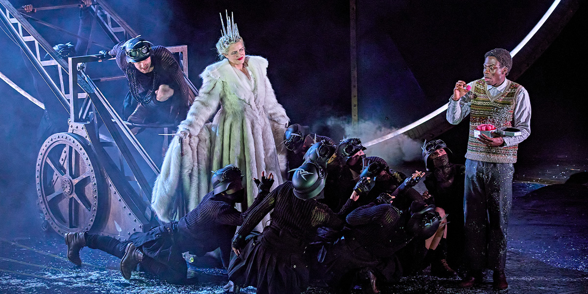 Edmund holds a box of Turkish Delight. The White Queen stands close by smiling while people wearing black clothing have fallen onto the floor.