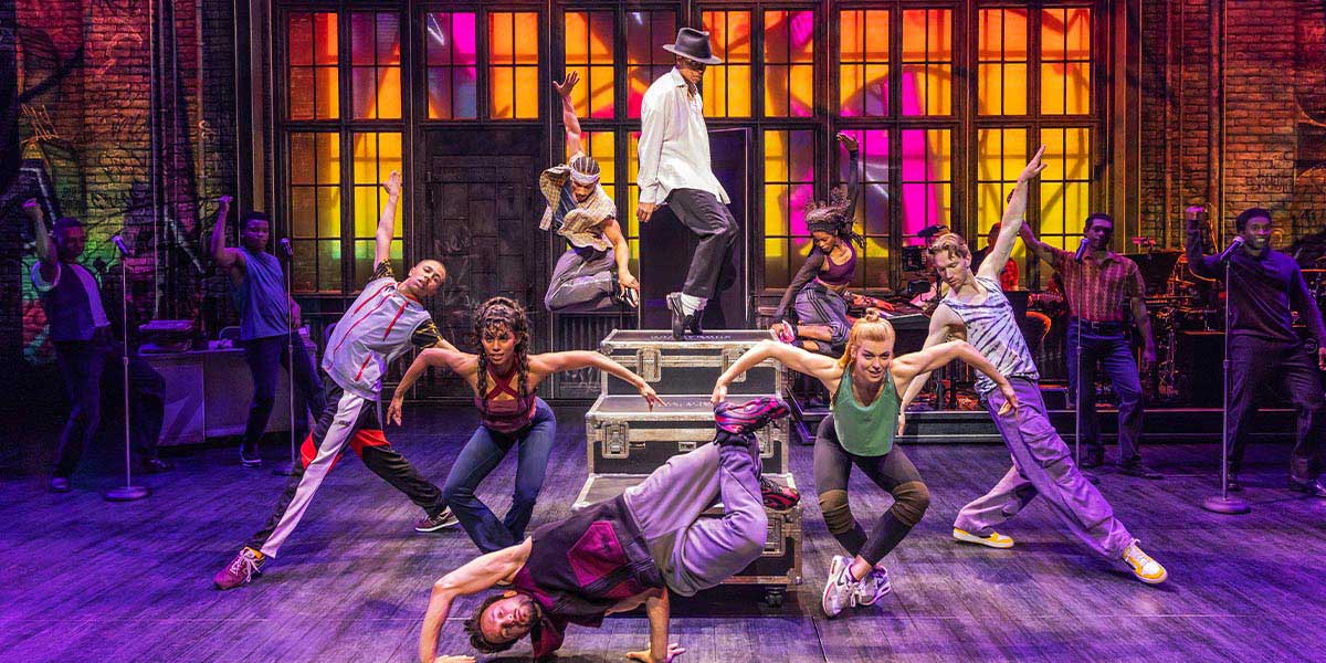 A group of performers in vibrant costumes poses energetically on stage during a scene from MJ The Musical.