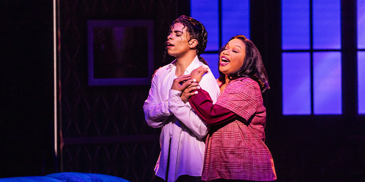 A performer in a vibrant costume strikes a dynamic pose on stage, with dramatic lighting and a cheering audience.