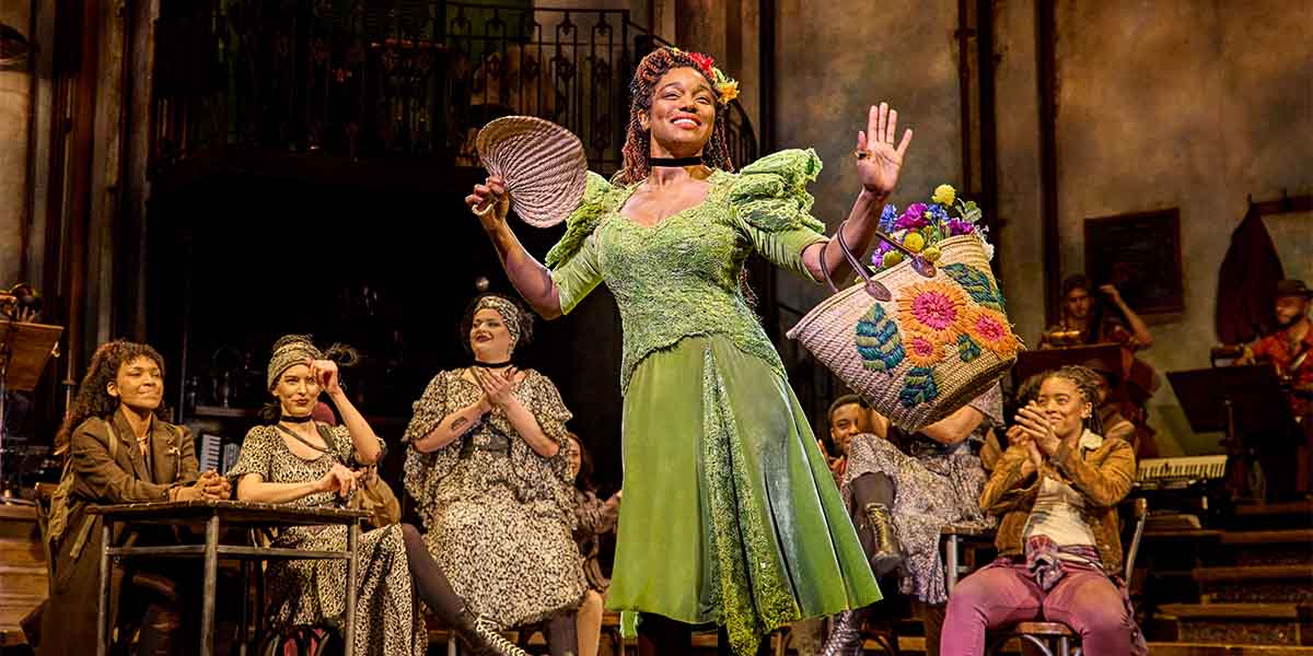 Rachel Adedeji and the Hadestown cast pose together, smiling, in vibrant costumes against a theatrical backdrop.