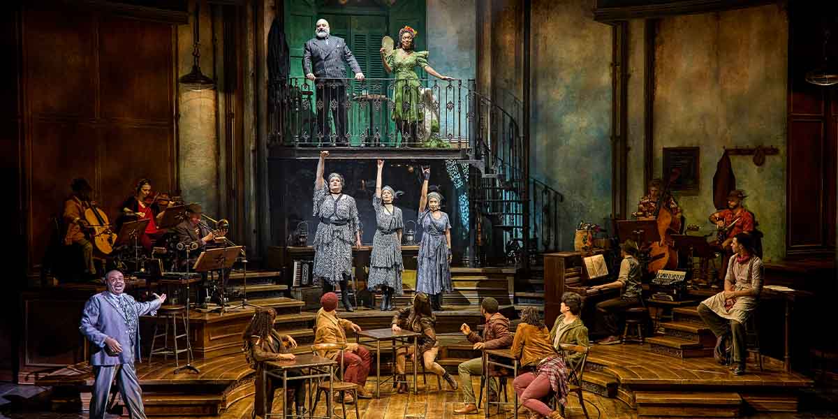 The cast of Hadestown performs on stage, featuring musicians and actors in vibrant costumes, with a detailed set.