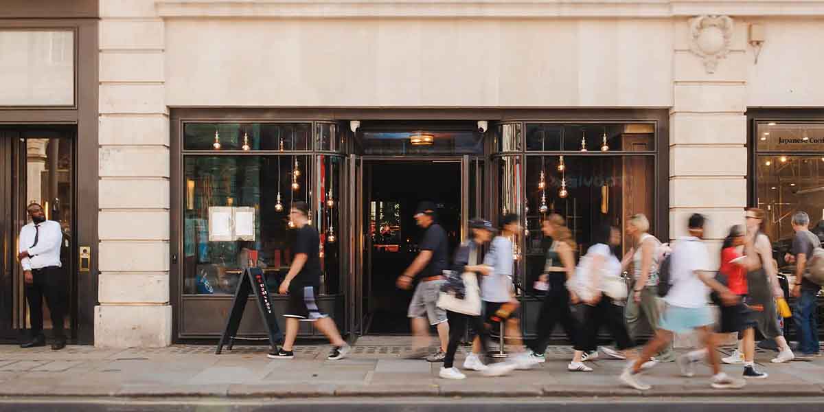 A diverse group of people walks past a restaurant entrance, with a doorman standing nearby.