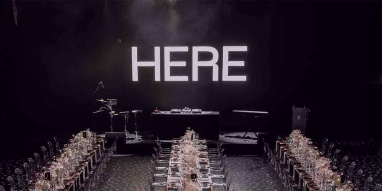 A large "HERE" sign illuminates a banquet setup with elegant tables and musical instruments in the background.
