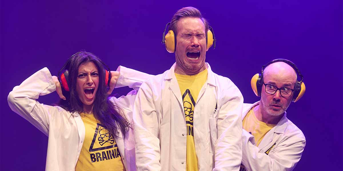 Three performers in lab coats and yellow T-shirts, one screaming, under purple stage lighting.