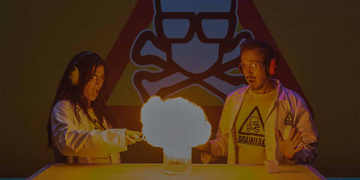 A woman in a lab coat ignites an experiment, creating a large flame, while a man reacts surprised.