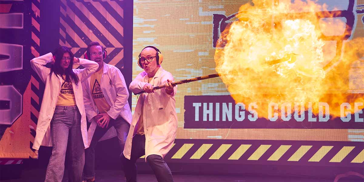 Three performers in lab coats, one igniting a fireball, with a vibrant, caution-themed background.