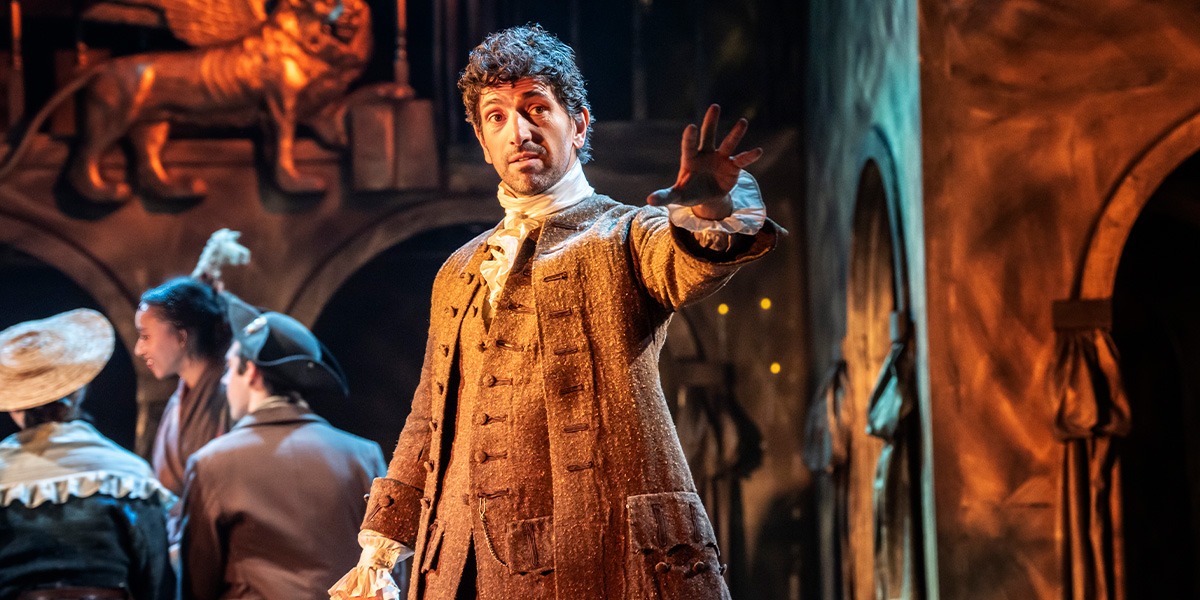 A person is standing holding out their right arm in front of them. They are wearing a pale brown textured suit with a white ruffled collar and cuffs.