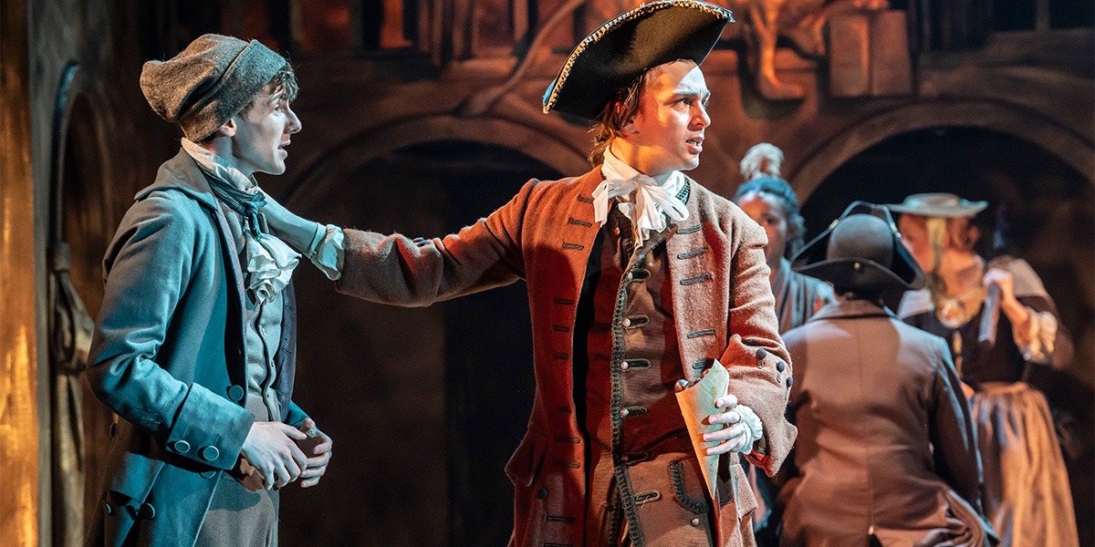 two people are standing beside one and other. The person on the left is wearing a grey beanie style hat, a grey suit with a white ruffled collar. The person on the right is wearing a brown suit with a white neck ribbon and a triangular black hat. The person on the left has their hand on the others shoulder.