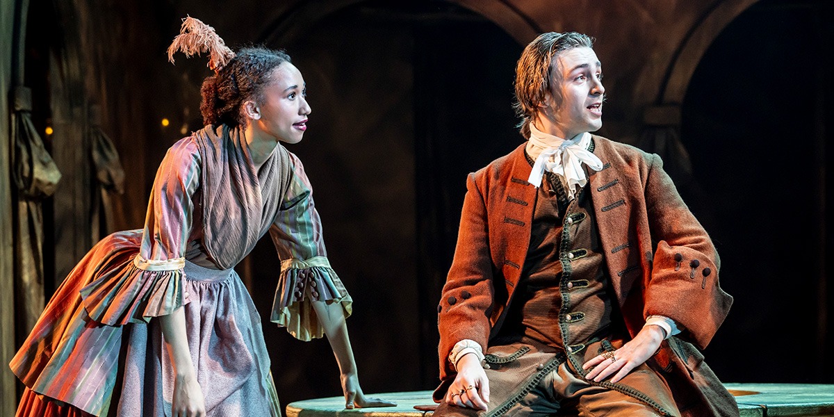 There is a person wearing a frilly multi-coloured dress they are standing beside another person who is wearing a brown suit with a white neck ribbon. They are sitting on a large, round, wooden table.
