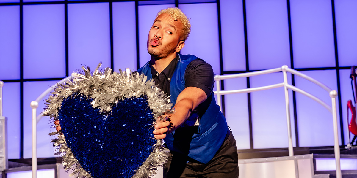 Danny Kaan poses confidently in a stylish outfit, with a blurred background featuring a theatre setting.