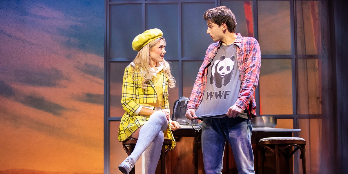 Emma Flynn (Cher) is sitting down wearing a yellow checked skirt suit and hat. Keelan McAuley (Josh) is standing beside wearing a WWF t-shirt.
