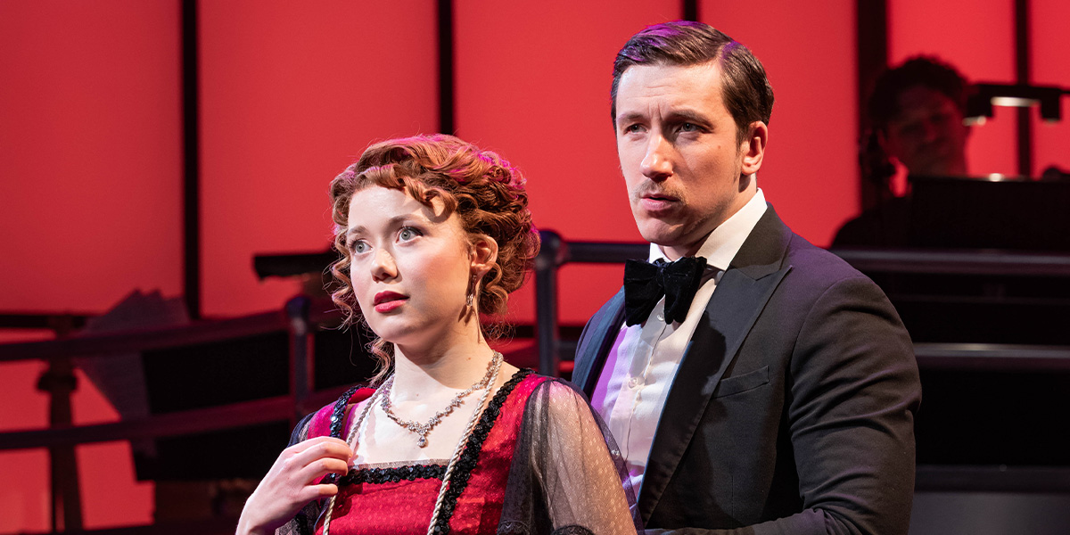 A senior man with glasses smiles, wearing a dark suit, against a blurred background of a theatre.