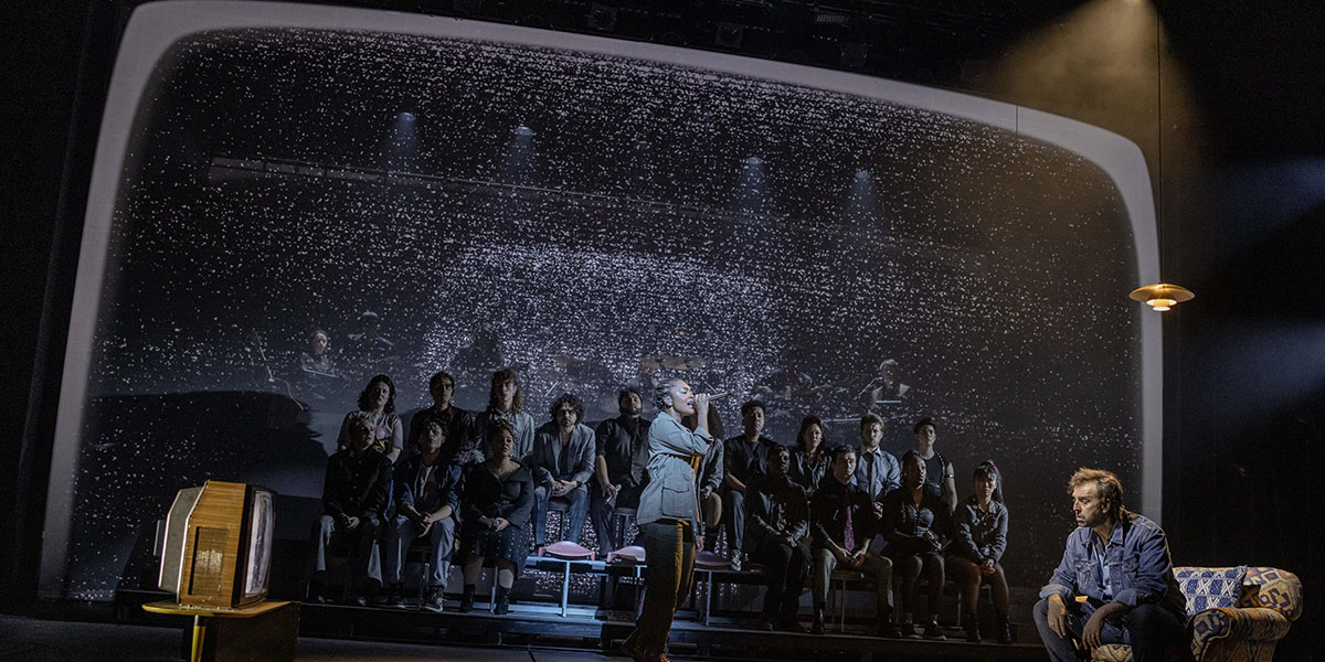 A performer in a vibrant costume sings passionately on stage, with colourful lights illuminating the background.