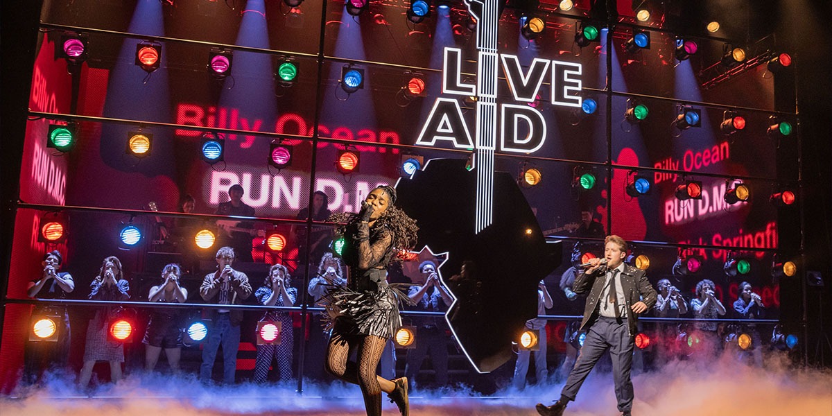 A performer in a vibrant costume sings passionately on stage, with colourful lights illuminating the background.