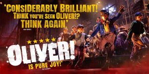 A theatrical scene from "Oliver!" featuring actors in period costumes, with a dimly lit stage backdrop.