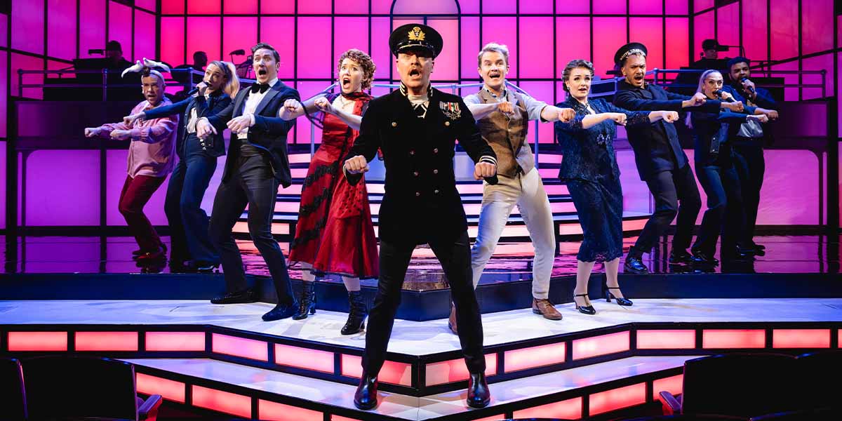 Tom Allen and the cast of Titanique pose together, wearing vibrant costumes, with a theatrical backdrop.