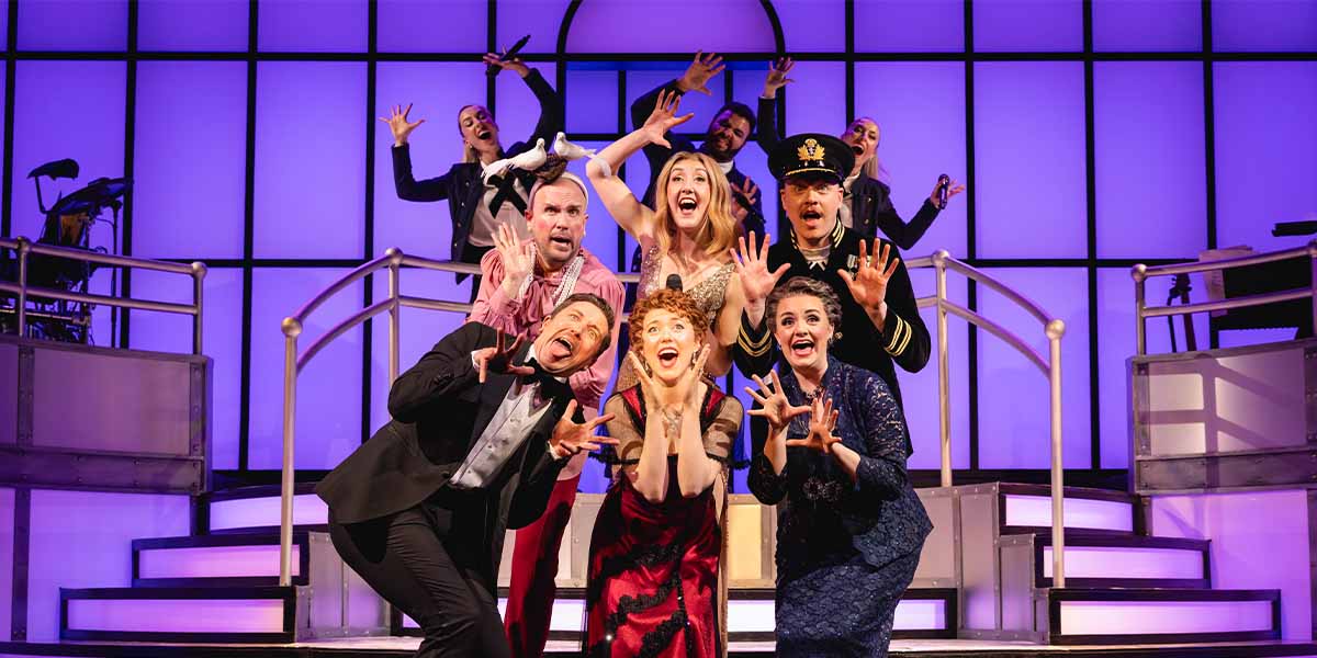 Tom Allen and the cast of Titanique pose together, smiling, wearing vibrant costumes against a theatre backdrop.