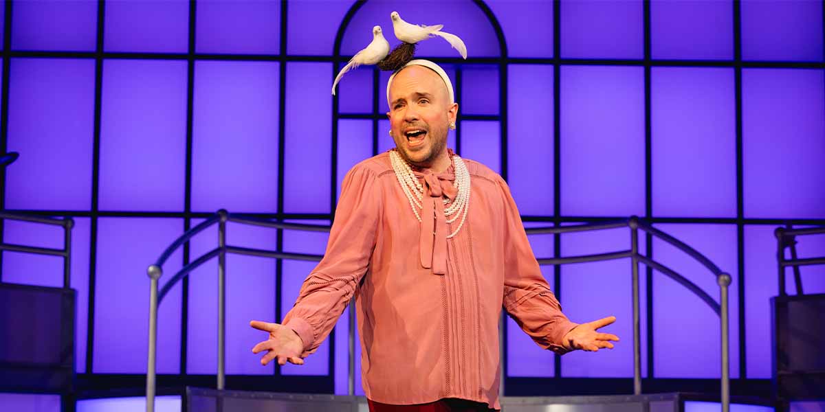 Tom Allen performs in a pink blouse with pearls, two white doves on his head, against a purple backdrop.